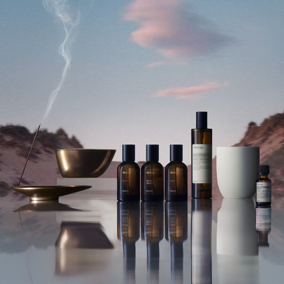 A collection of Aesop products, including perfumes, room spray, and essential oil, with an incense stick burning in a bronze holder, set against a blurred mountainous landscape reflected on a surface
